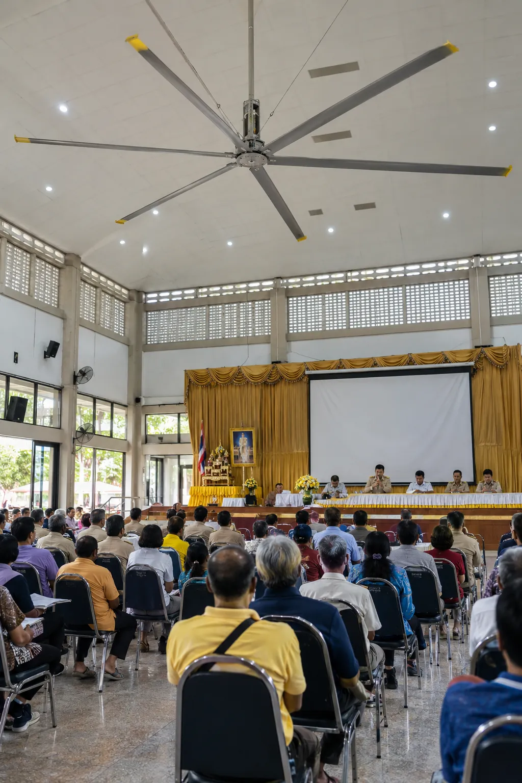 พัดลมยักษ์ใต้เพดานหอประชุมของหน่วยงานราชการ ผู้เข้าประชุมนั่งฟังหน้าเวที