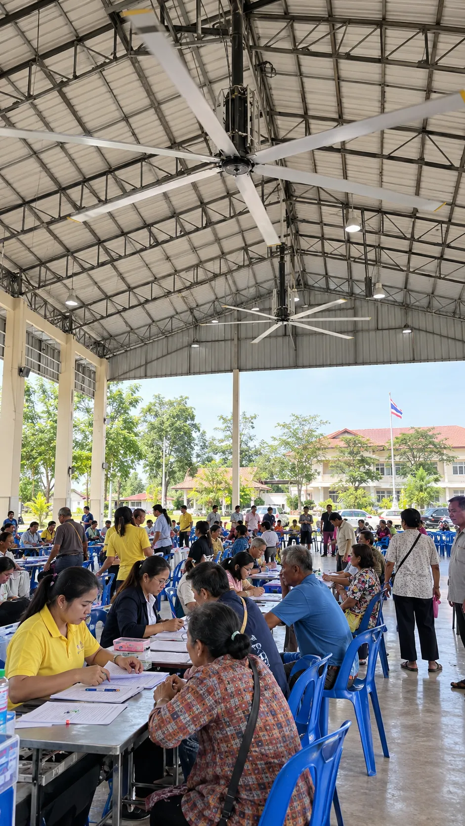 พัดลมยักษ์ใต้โครงหลังคาเหล็กของศาลาอเนกประสงค์ เจ้าหน้าที่ให้บริการประชาชนที่โต๊ะลงทะเบียน