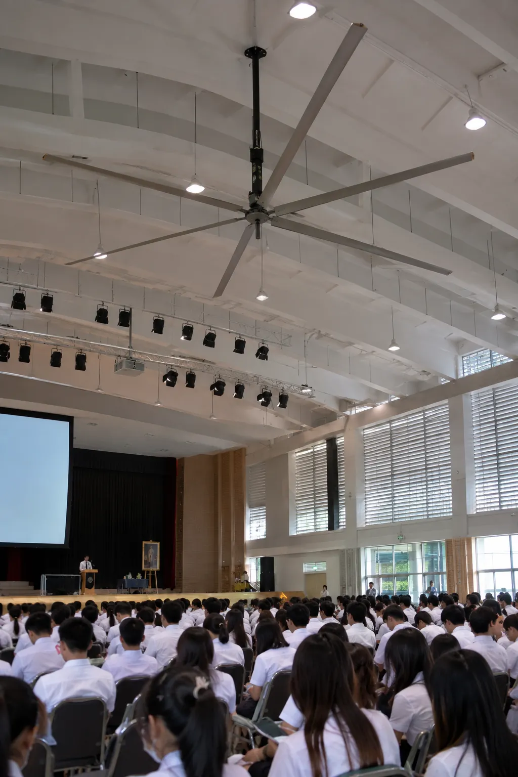 พัดลมยักษ์ใต้เพดานหอประชุมโรงเรียน นักเรียนนั่งฟังกิจกรรมหน้าเวที