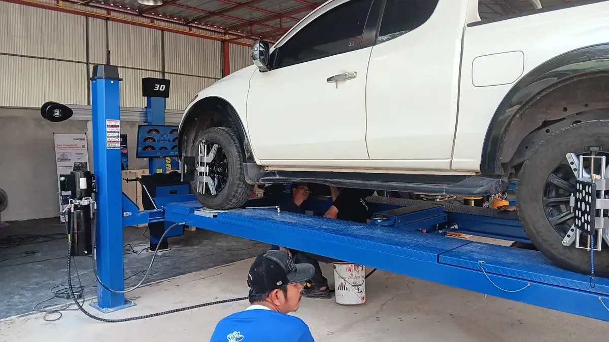 รถกระบะบนลิฟท์ 4 เสาตั้งศูนย์ Mactech สีน้ำเงิน ติดหัวจับล้อและเป้าวัด หน้าเสากล้องเครื่องตั้งศูนย์ 3D ช่างกำลังปรับช่วงล่างใต้รถ